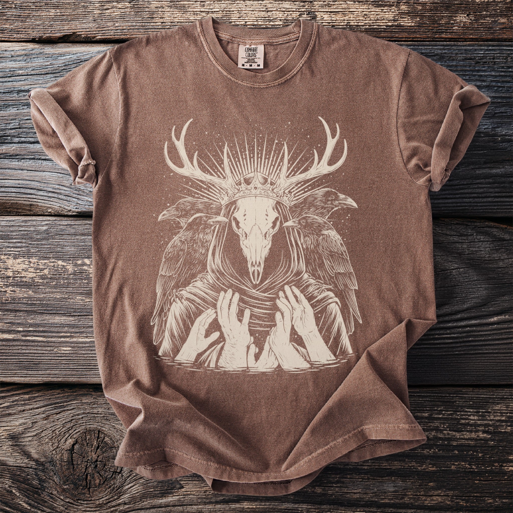 Brown t-shirt with depiction of a crowned deer skull with ravens and hands.