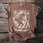Brown t-shirt featuring a graphic of a creature carrying bones and a staff with snowflakes and trees.