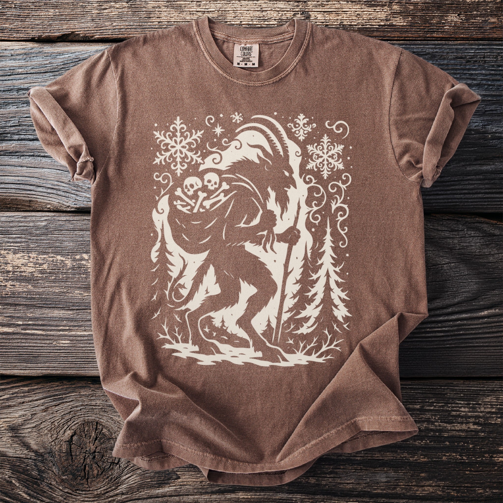 Brown t-shirt featuring a graphic of a creature carrying bones and a staff with snowflakes and trees.