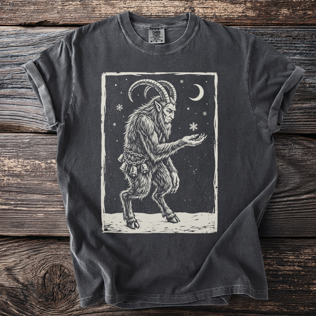 Pepper gray t-shirt with a cream folk-cut print: Krampus stands in the snow, chains and bells on, holding out one hand for a falling snowflake.