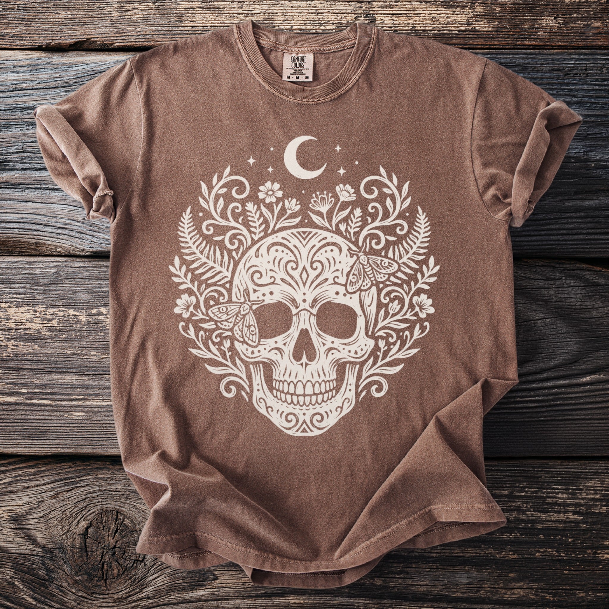 Brown t-shirt with white skull, floral, and moth design and moon.