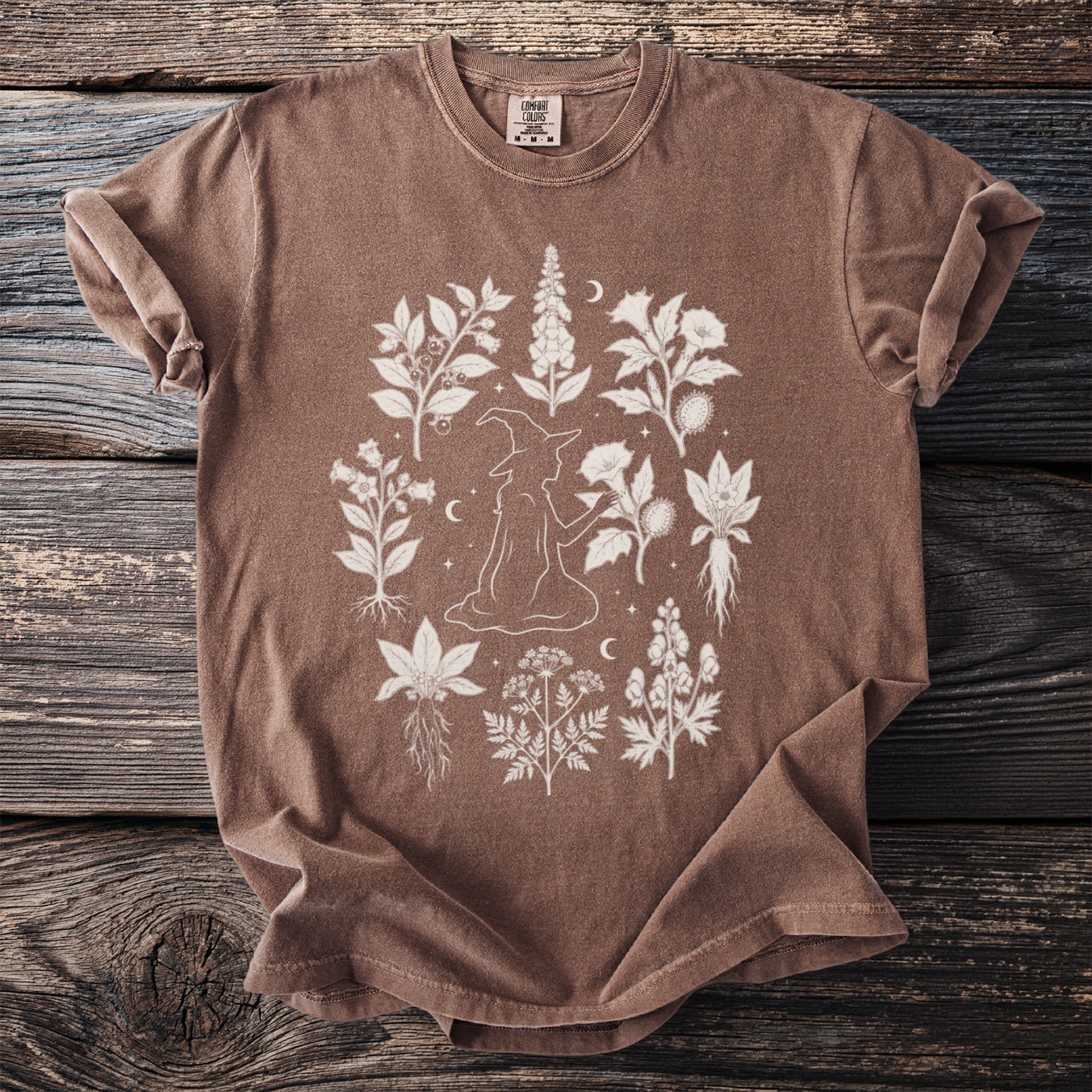 Brown t-shirt with white linework drawing of a witch surrounded by various plants.