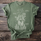 Green t-shirt with white line art of a witch holding a mortar and pestle, surrounded by plants and text.