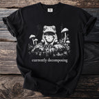 Black t-shirt graphic of a tired frog sitting among mushrooms with text that reads "currently decomposing".
