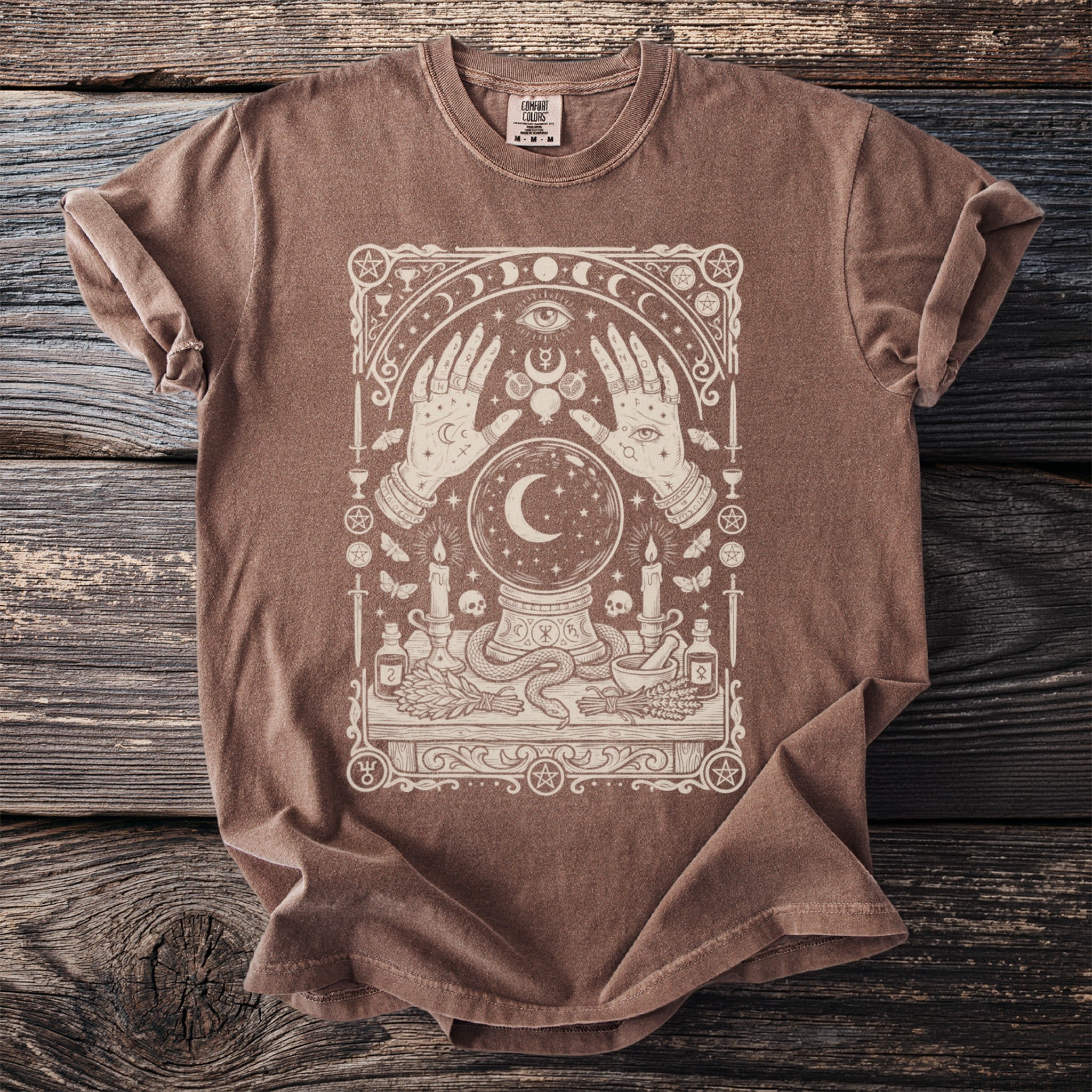 Brown t-shirt with cream-colored mystical graphic featuring hands, crystal ball, moon, and celestial elements.