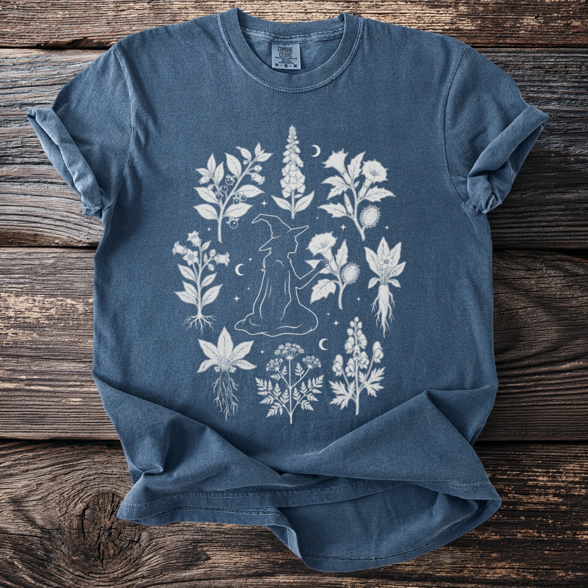 Blue t-shirt with a white line drawing of a witch holding a flower surrounded by botanical elements.
