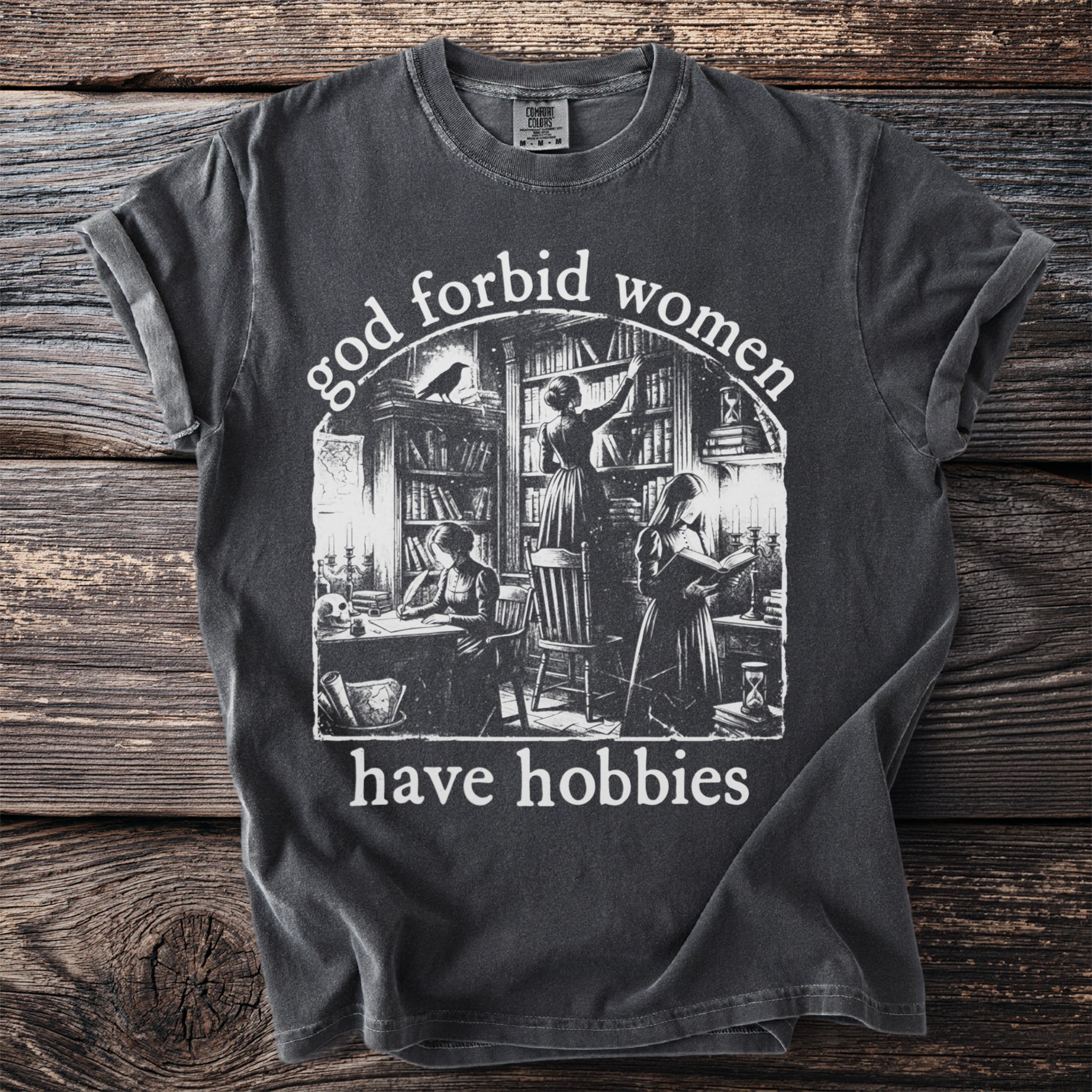 Dark grey t-shirt with "god forbid women have hobbies" text and library illustration.