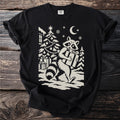 Black t-shirt with a cream folk-cut print: a raccoon walks away from a decorated tree in the snow, a wrapped present under one arm.