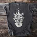 Dark t-shirt with a cream graphic of a witch surrounded by plants and a crescent moon.