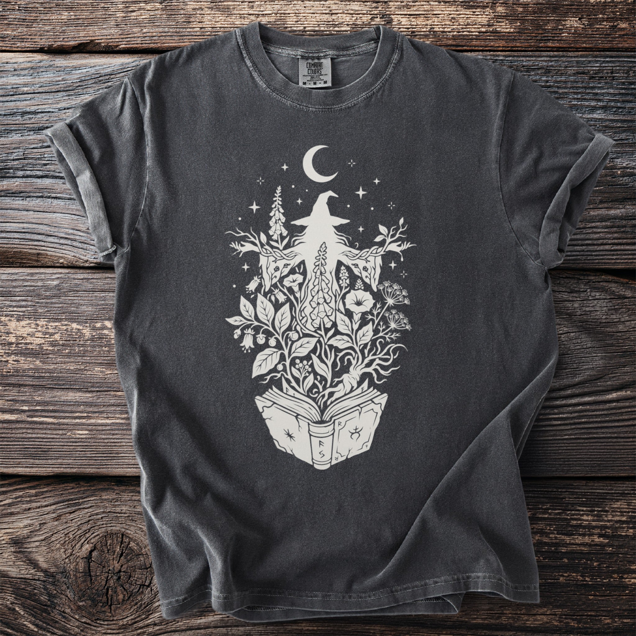 Dark t-shirt with a cream graphic of a witch surrounded by plants and a crescent moon.