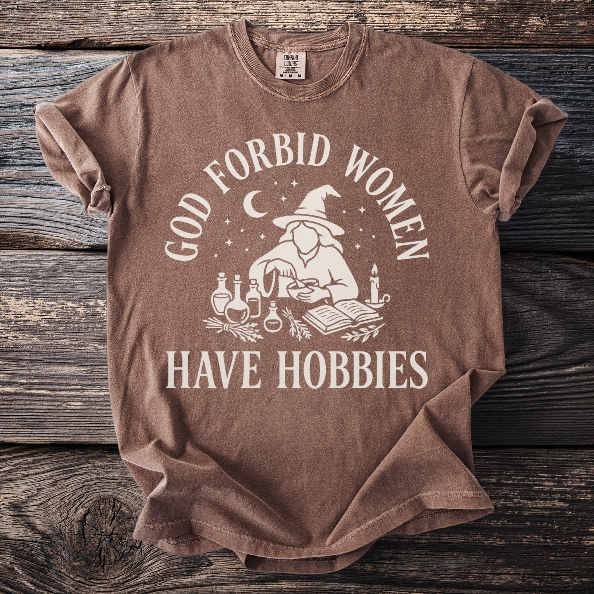 Brown t-shirt with cream graphic design that reads "God forbid women have hobbies."