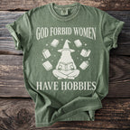 Green t-shirt with white text "GOD FORBID WOMEN HAVE HOBBIES" and witch reading a book surrounded by books.