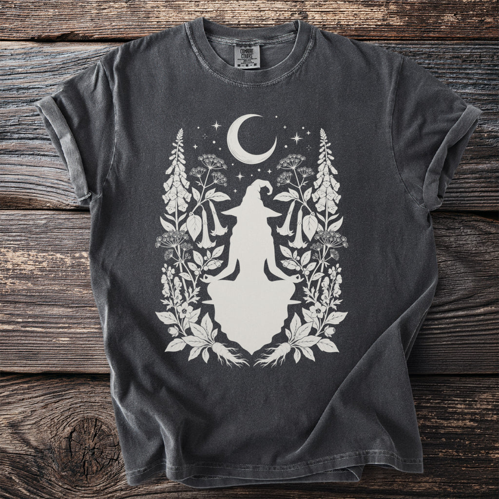 Dark t-shirt with a white silhouette of a meditating witch, moon, stars, and botanical elements.
