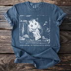 Blue t-shirt with a black and white graphic of an opossum eating from a wrapper surrounded by trash.