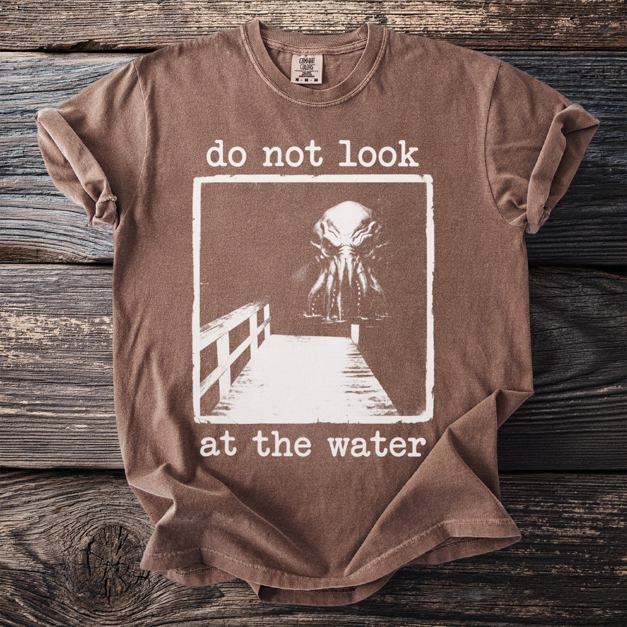 Brown t-shirt with "do not look at the water" and a creature on a dock.