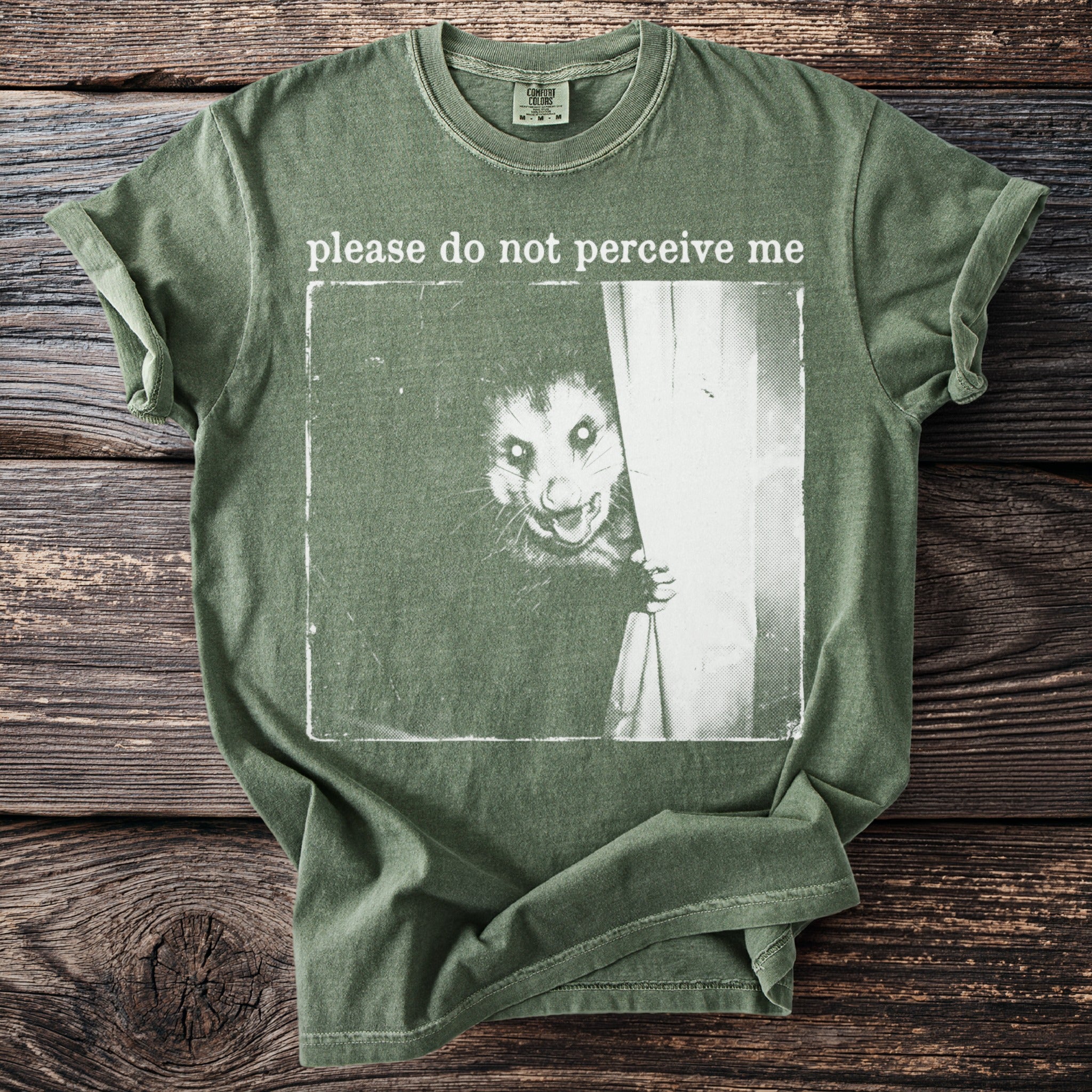 Green t-shirt with a vintage style graphic of a possum behind a curtain and text "please do not perceive me".