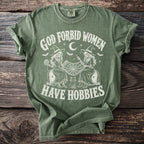 Green t-shirt with "God Forbid Women Have Hobbies" text and witch skeletons knitting.