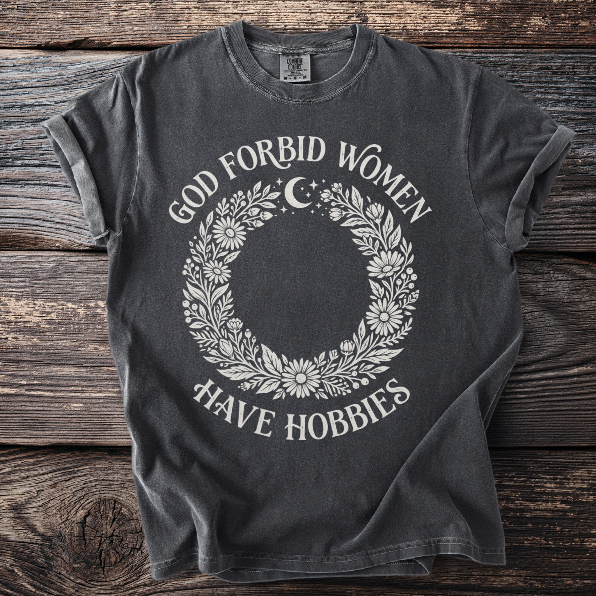 Dark grey t-shirt with "God Forbid Women Have Hobbies" text and floral moon design.