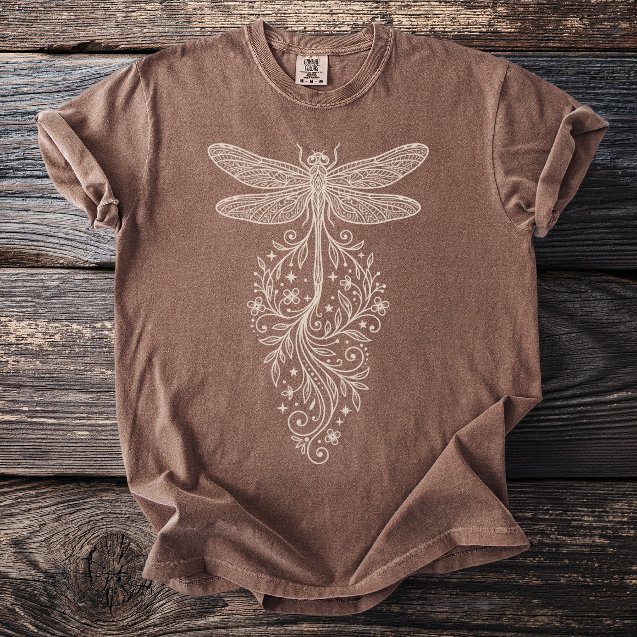 Brown t-shirt with cream-colored dragonfly and floral graphic on distressed wood.