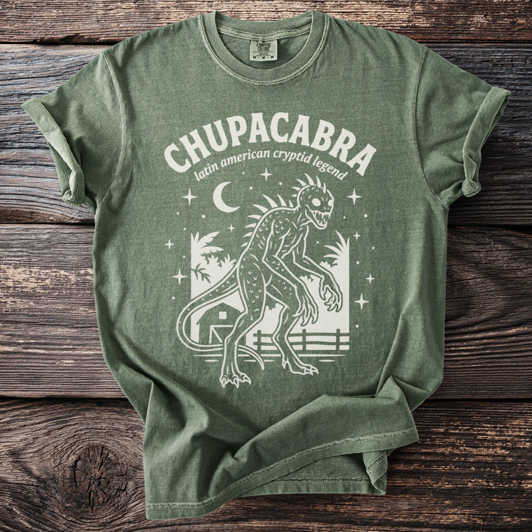 Green t-shirt with white graphic of a Chupacabra, moon, stars, and a barn.