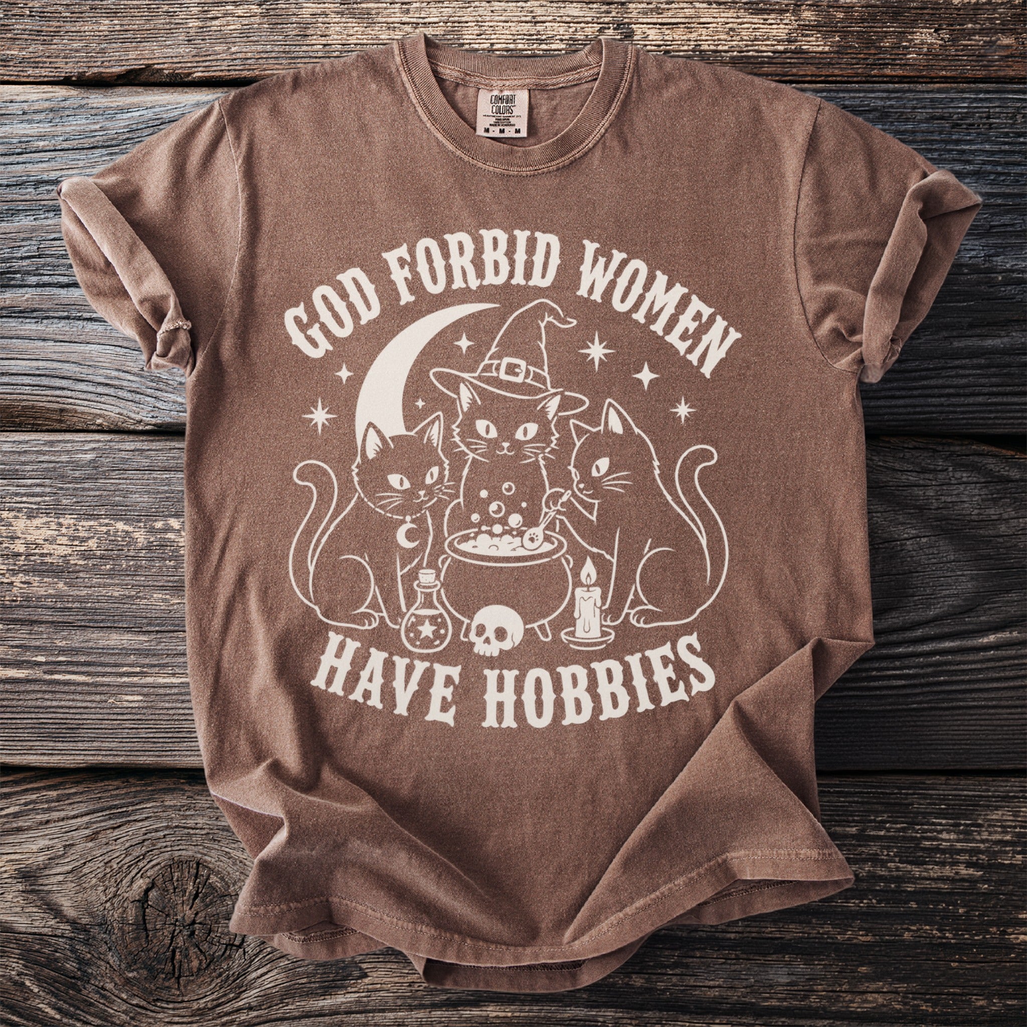 Brown shirt with white graphic of cats, cauldron, moon, and text "GOD FORBID WOMEN HAVE HOBBIES".
