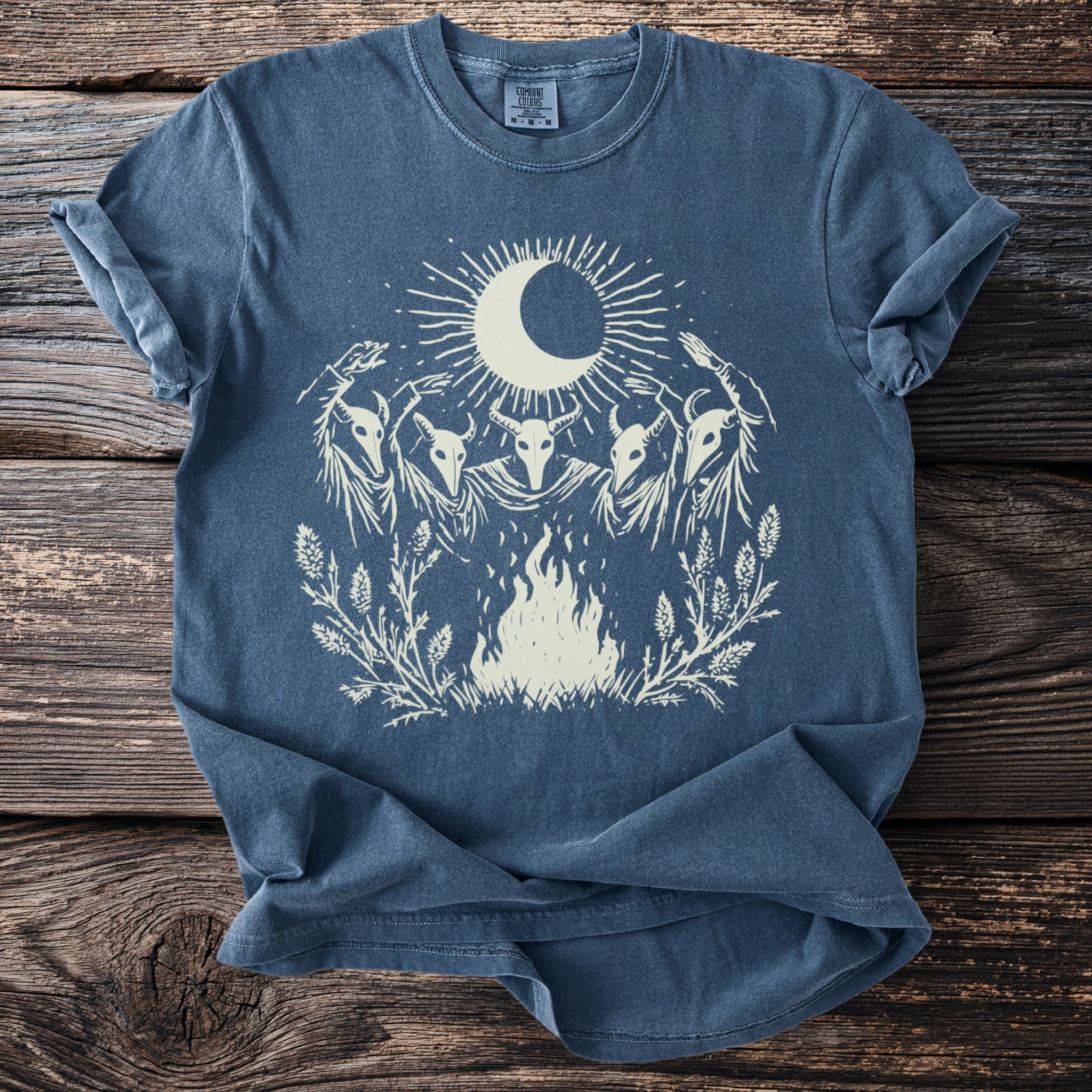 Blue t-shirt with white graphic of moon, sun, four skull figures, and campfire.