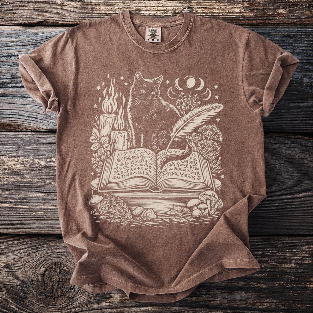 Brown t-shirt with cream print of a cat, open book, candles, and celestial elements.