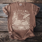 Brown t-shirt with cream print of a cat, open book, candles, and celestial elements.