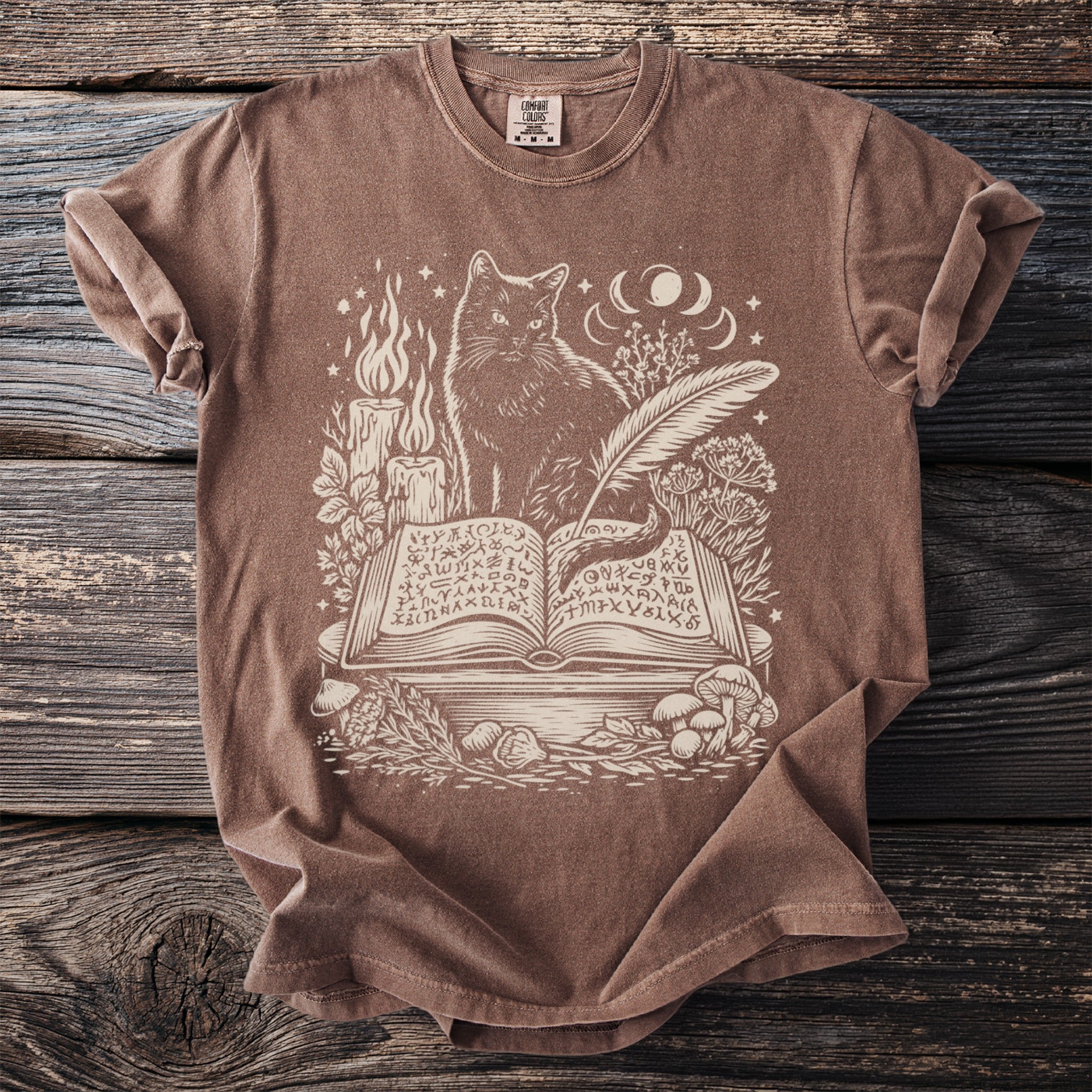Brown t-shirt with cream print of a cat, open book, candles, and celestial elements.