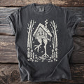 T-shirt with a cream graphic of a hut on chicken legs in a forest.