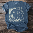 Blue Comfort Colors shirt with a vintage illustration of a Bigfoot-like creature in a forest under a full moon.