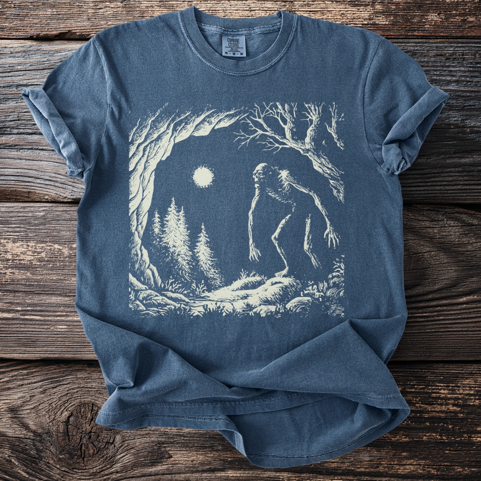 Blue Comfort Colors shirt with a vintage illustration of a Bigfoot-like creature in a forest under a full moon.
