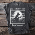 Dark t-shirt with graphic of witch in field and text "god forbid women have hobbies."