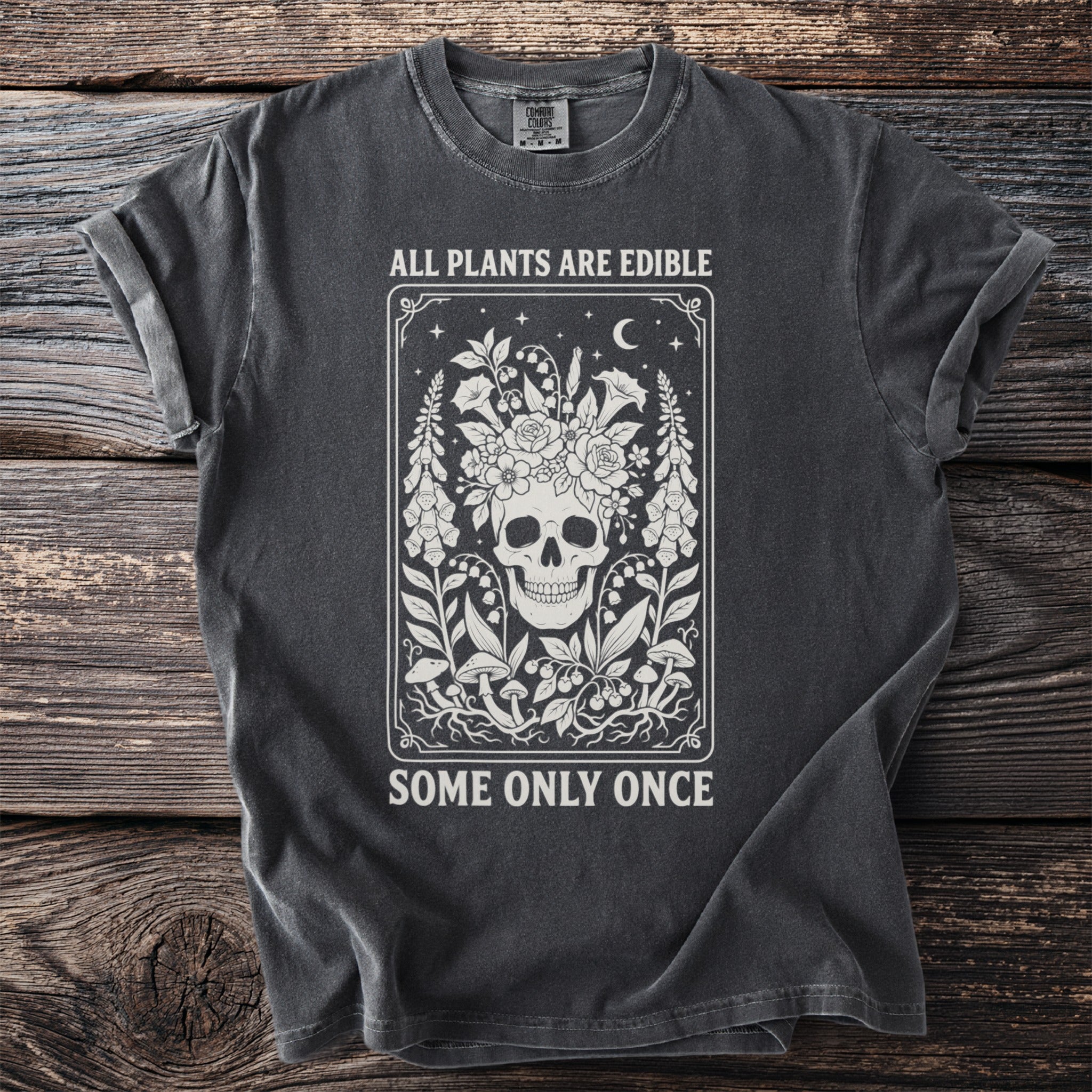 Dark grey t-shirt with skull, flowers, mushrooms, and text "ALL PLANTS ARE EDIBLE SOME ONLY ONCE".