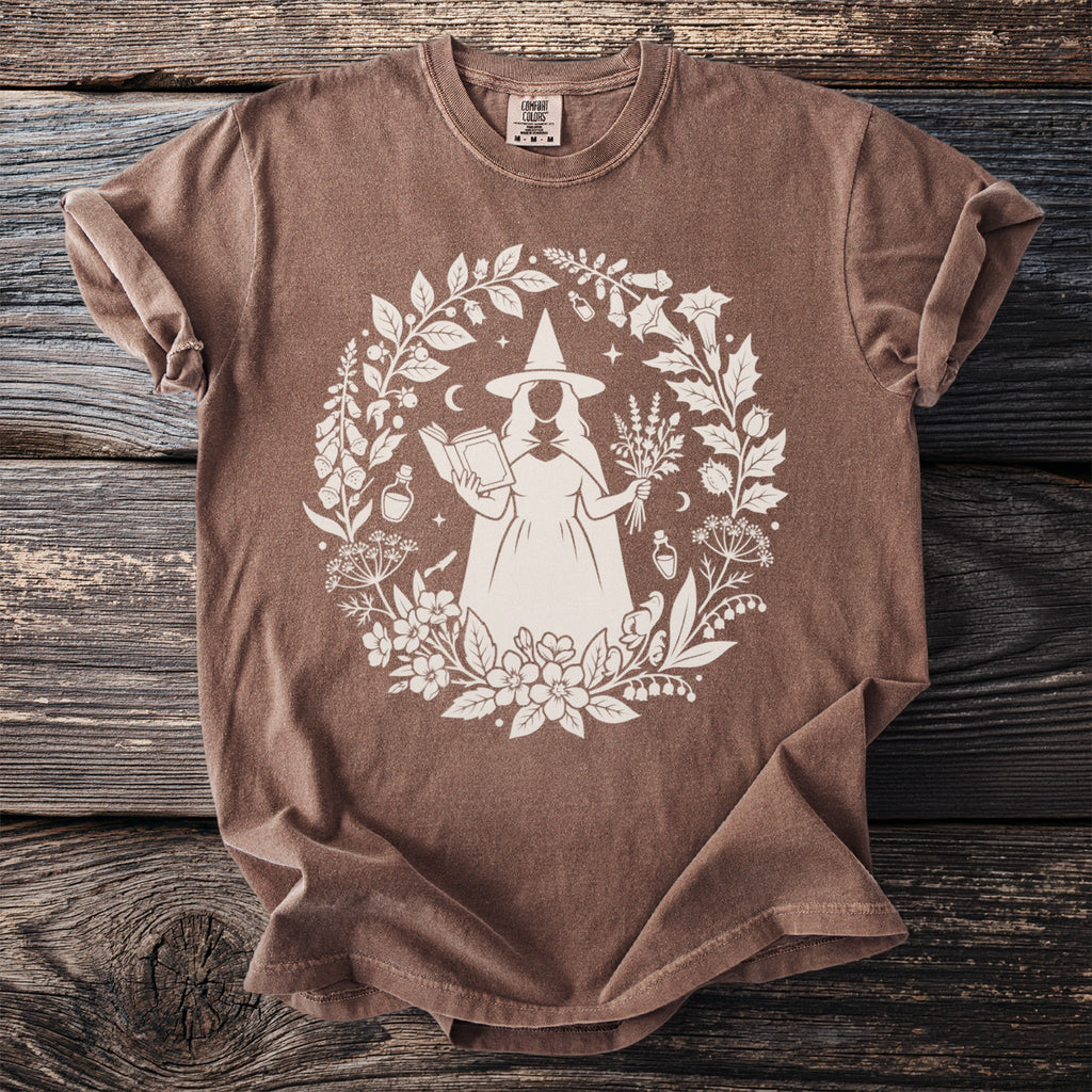 Brown t-shirt with a white circular print of a witch holding a book and herbs.