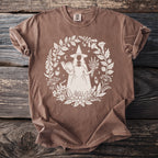 Brown t-shirt with a white circular print of a witch holding a book and herbs.
