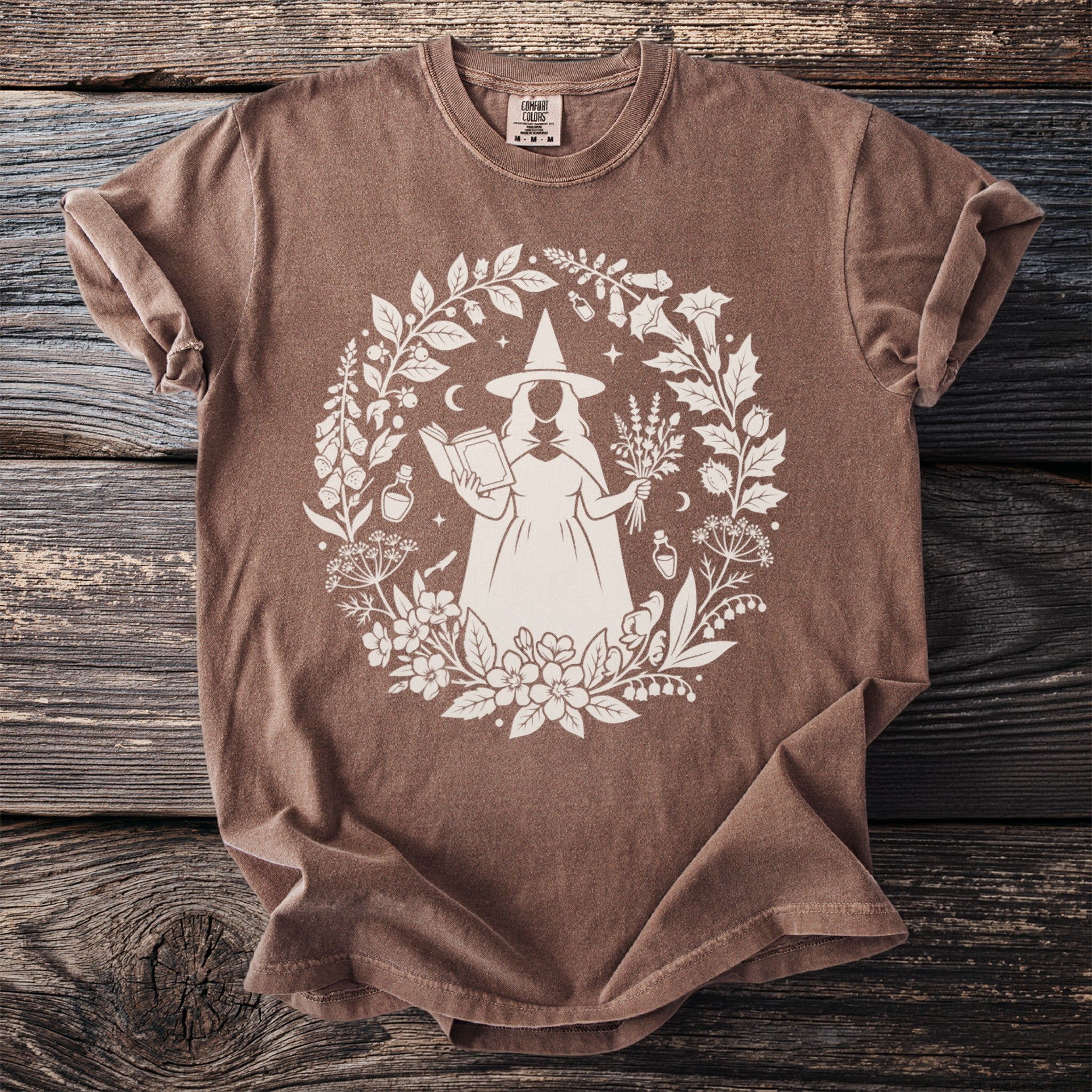Brown t-shirt with a white circular print of a witch holding a book and herbs.
