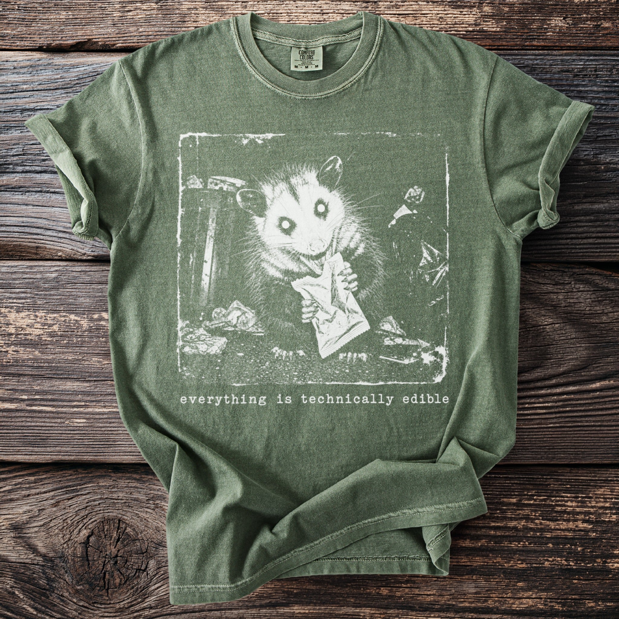 Green t-shirt with white print of a possum eating from a bag, saying "everything is technically edible".