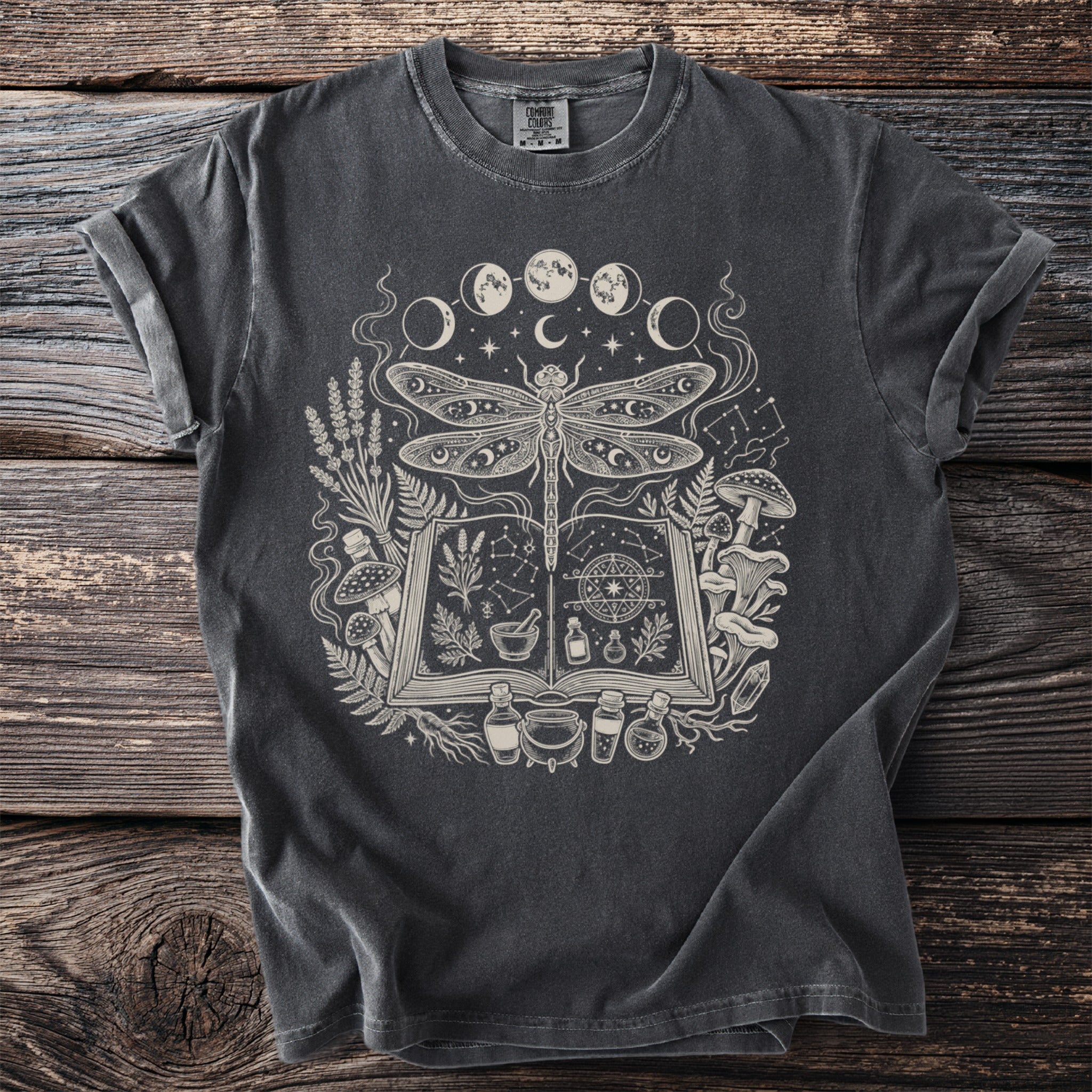 Dark gray t-shirt with a cream-colored print of a dragonfly, open book, moon phases, and mystical elements.