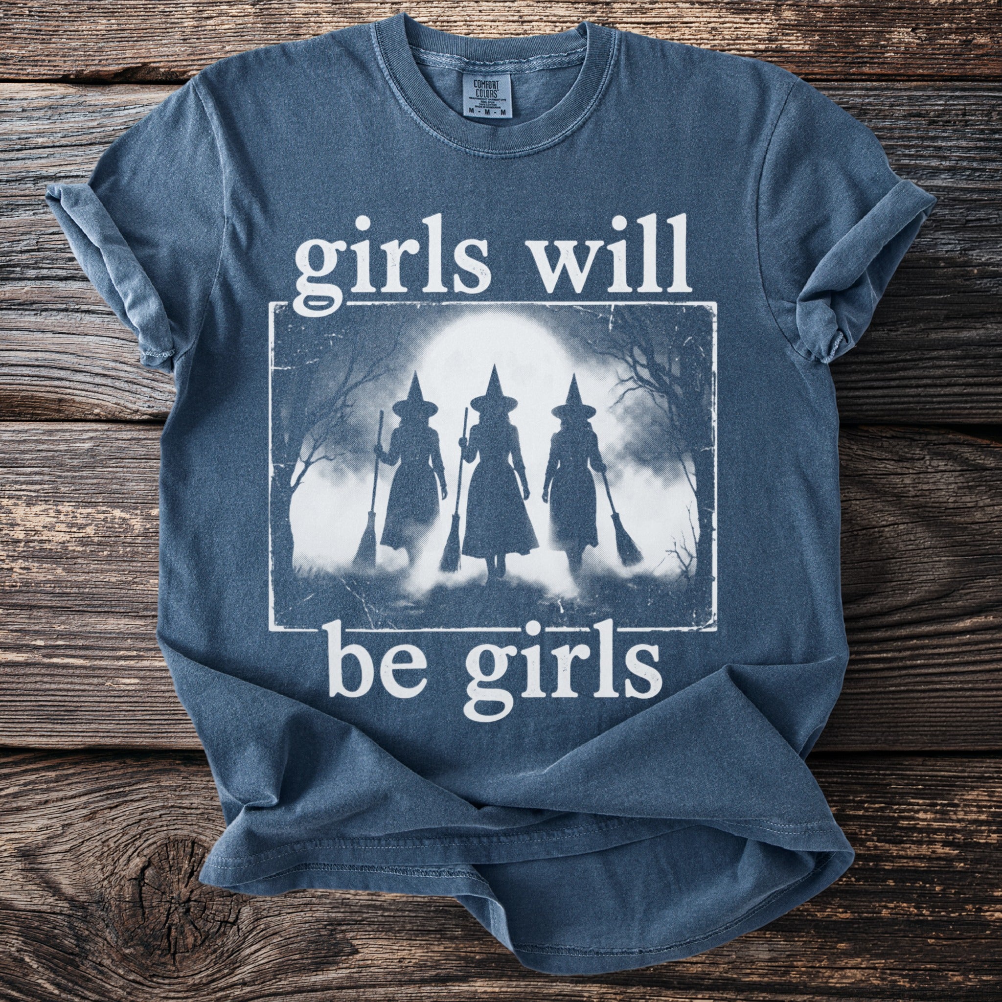 Blue t-shirt with white text "girls will be girls" and a graphic of three witches.