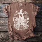 Brown t-shirt with a white line art graphic of two figures under a witch hat, surrounded by herbs and potions.