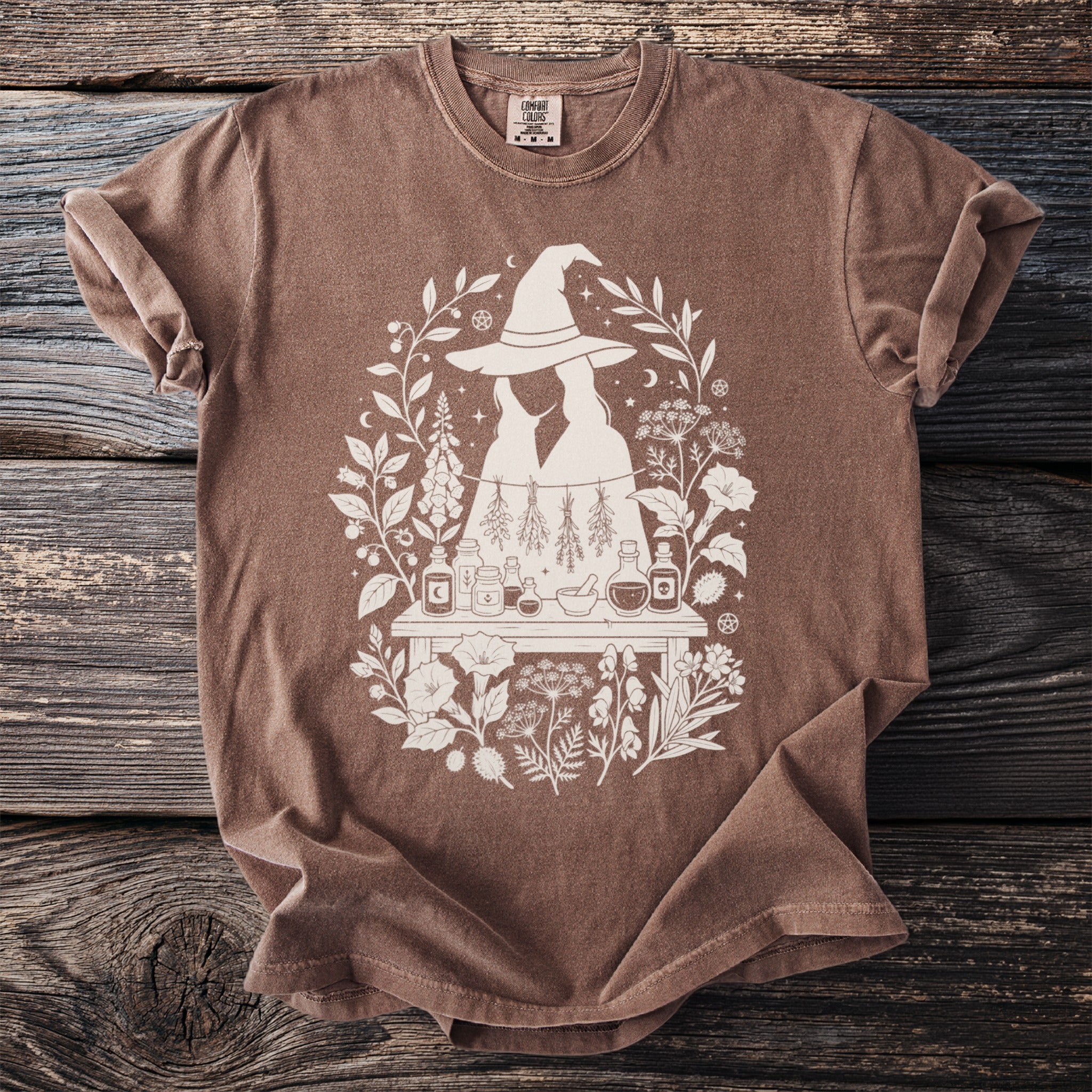 Brown t-shirt with a white line art graphic of two figures under a witch hat, surrounded by herbs and potions.
