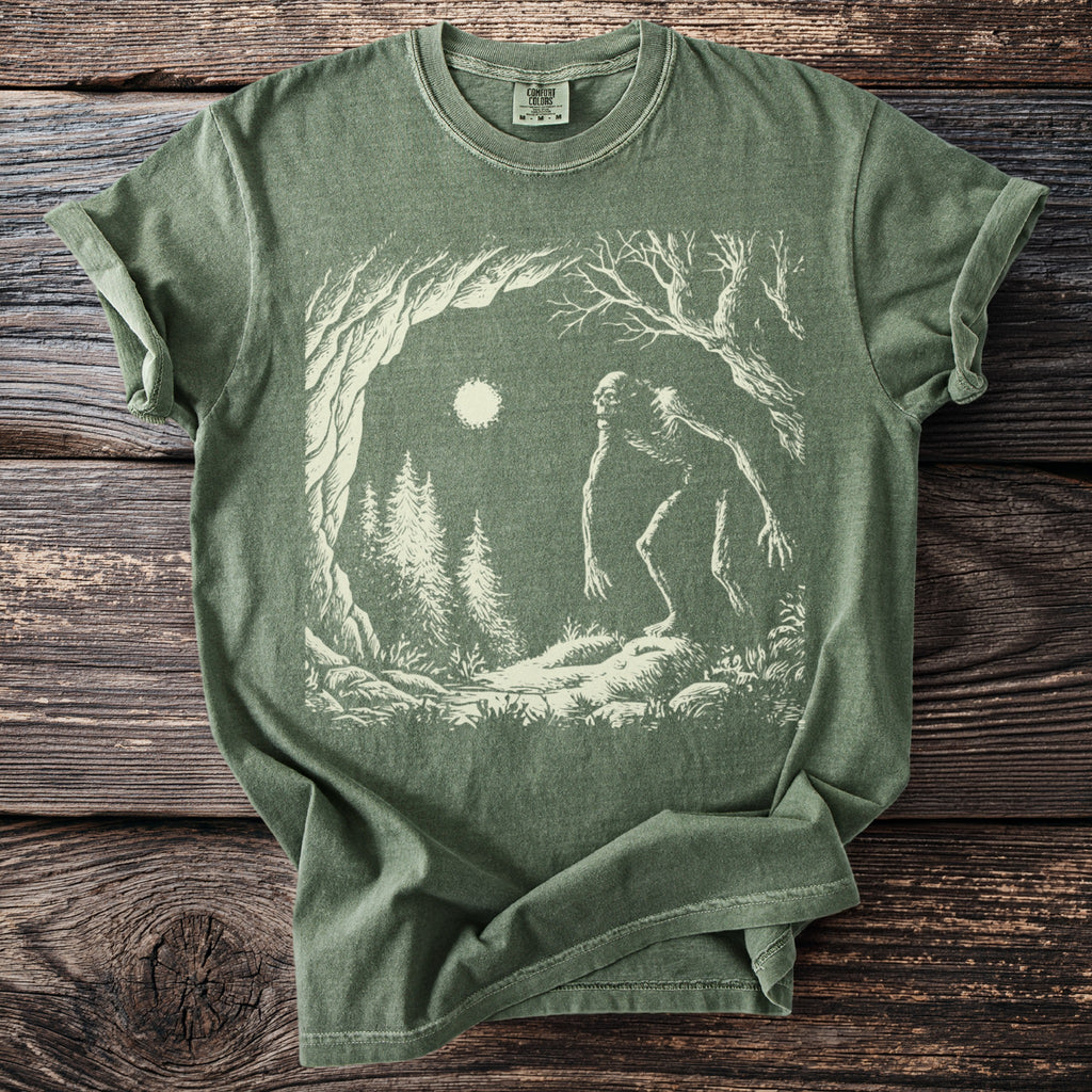 Green t-shirt with a cream graphic of a tall, thin creature in a forest at night.
