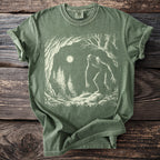Green t-shirt with a cream graphic of a tall, thin creature in a forest at night.