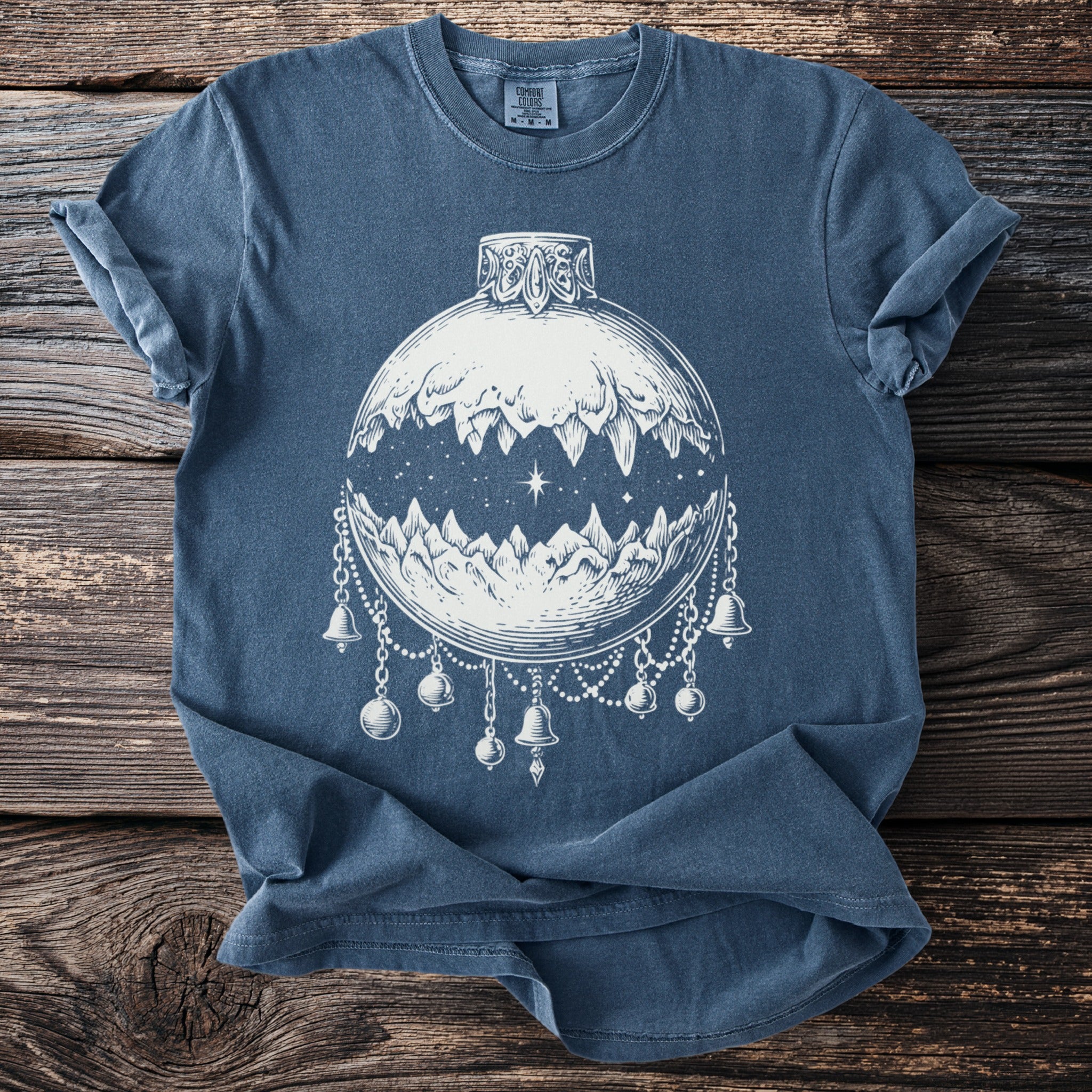 Blue t-shirt with white line art of a moon as a landscape containing mountains with bells and ornaments.