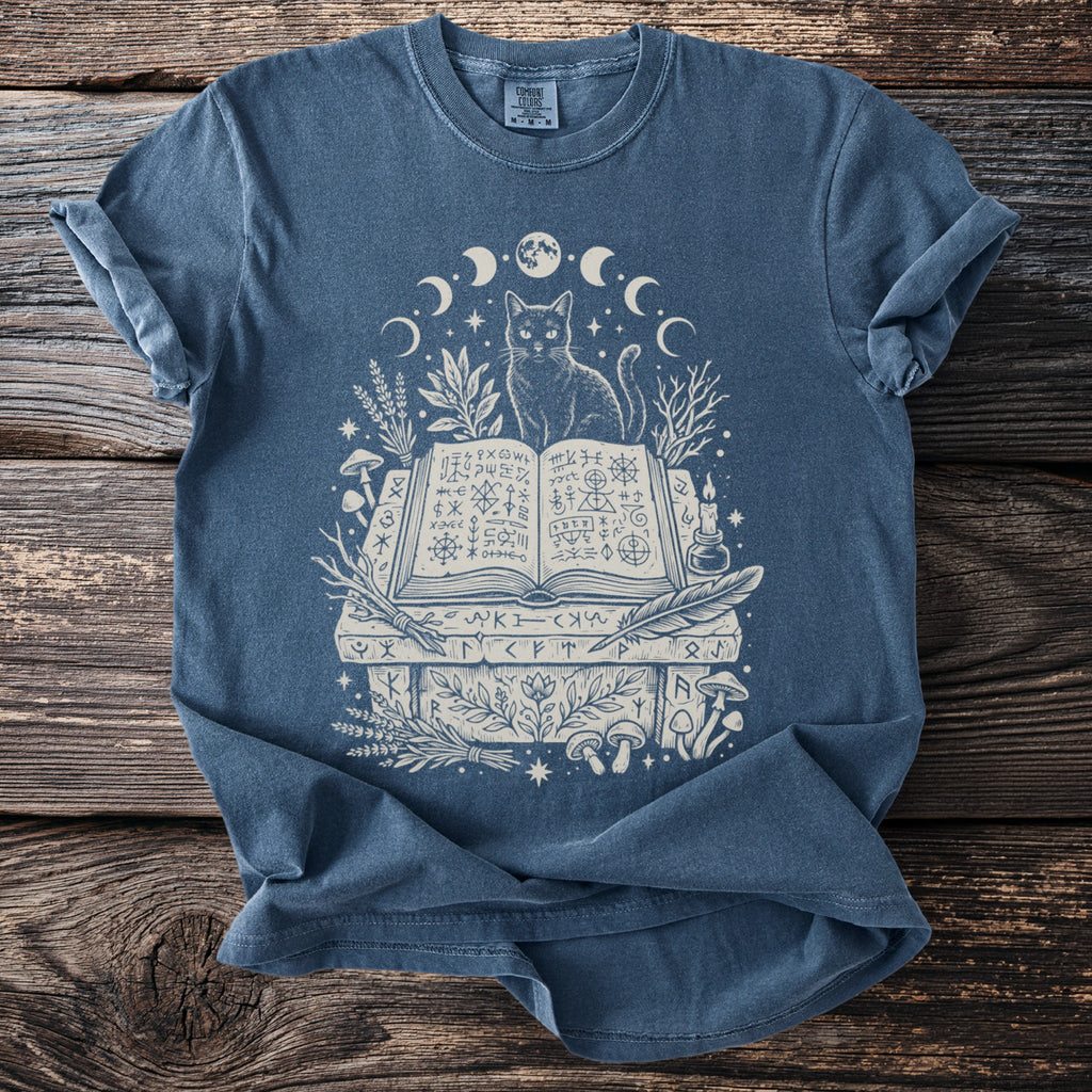 Blue t-shirt with a cat sitting on an open book surrounded by moons and mystical symbols.