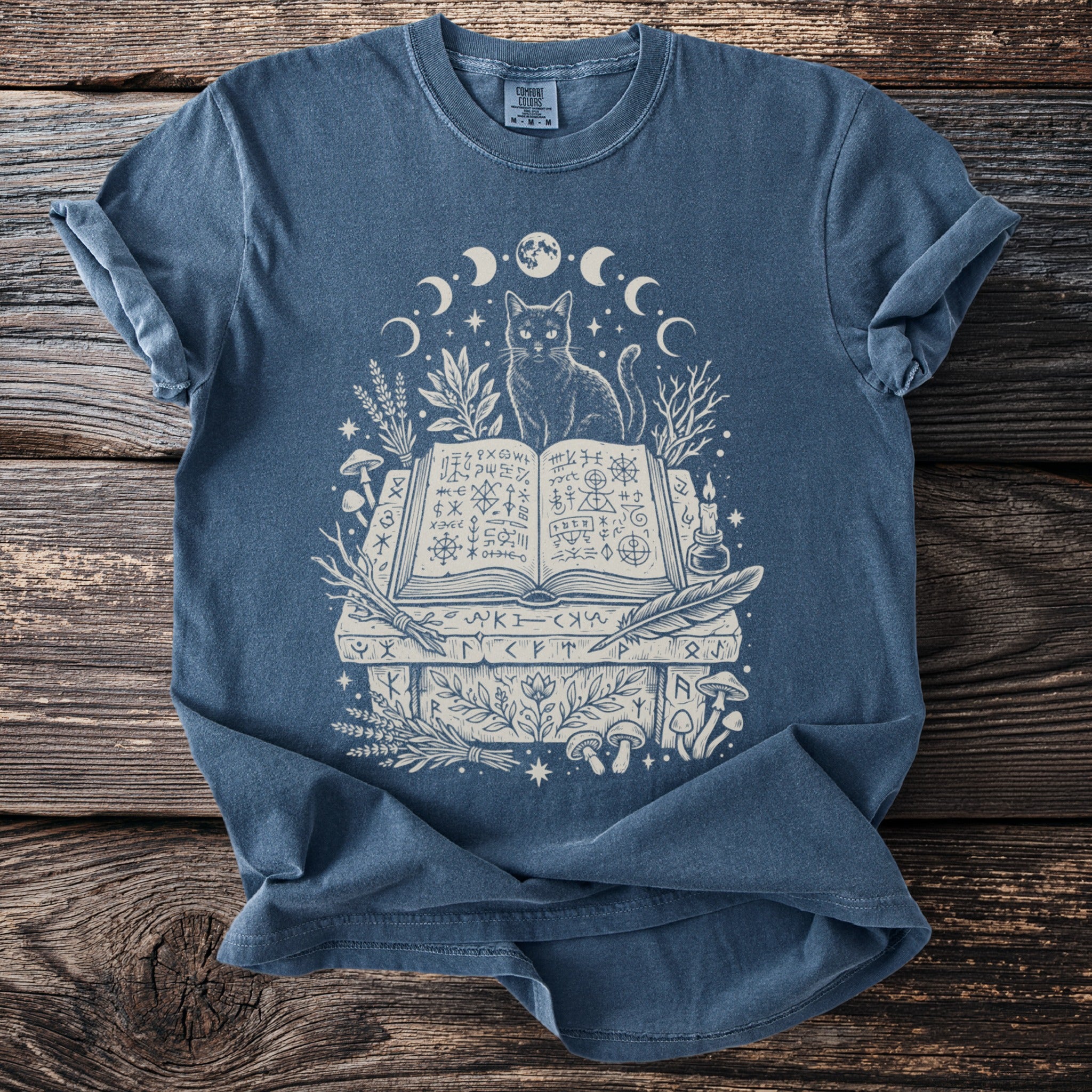Blue t-shirt with a cat sitting on an open book surrounded by moons and mystical symbols.