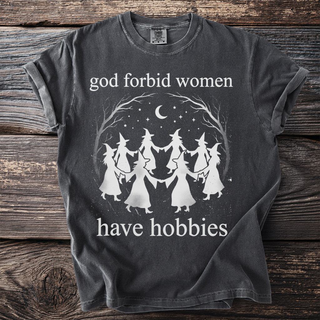 Dark t-shirt with "God forbid women have hobbies" text and witches dancing under a moon.