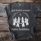 Dark t-shirt with "God forbid women have hobbies" text and witches dancing under a moon.