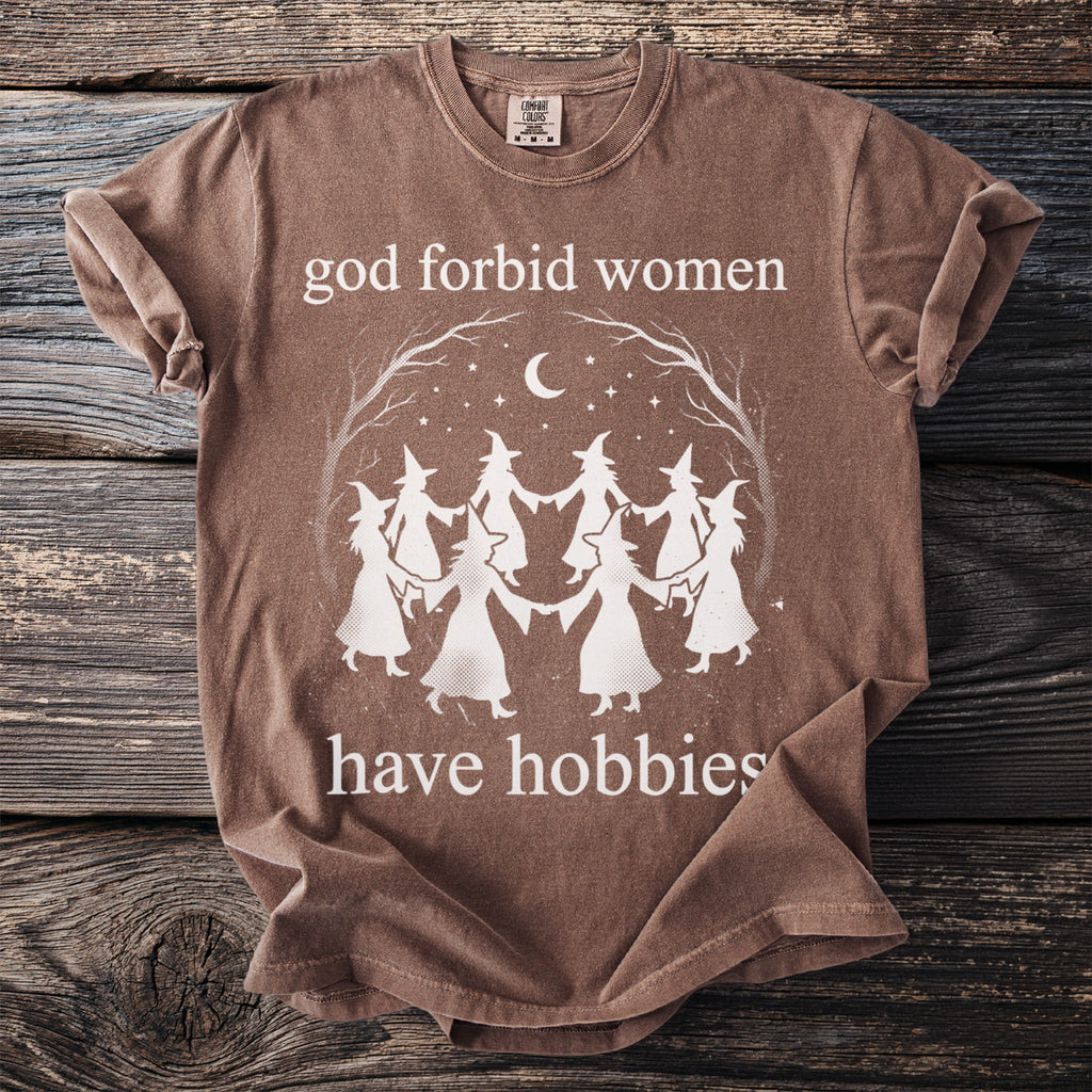Brown t-shirt with white text that reads "god forbid women have hobbies" and a graphic of witches dancing.